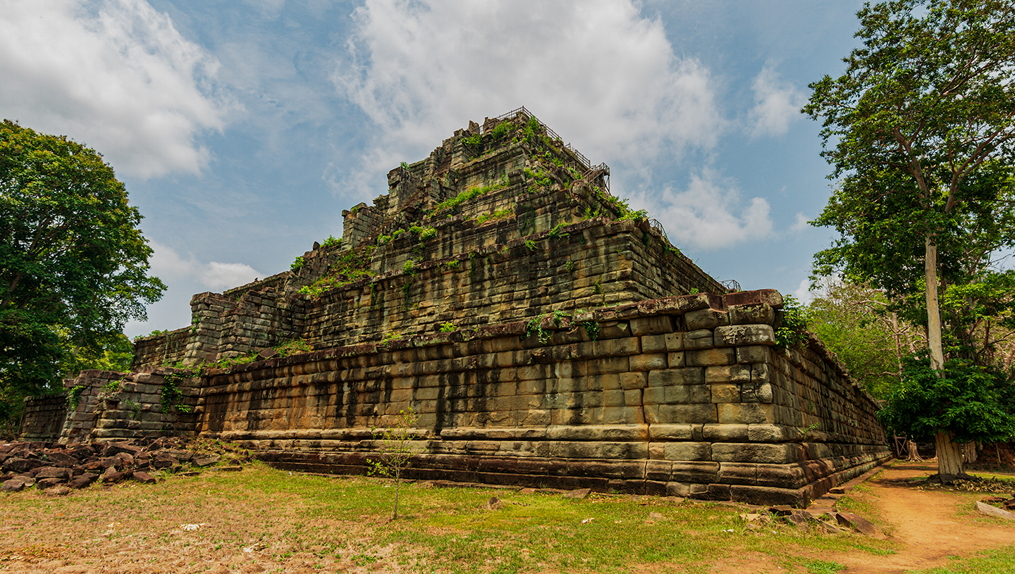 Zřícenina kamenné pyramidy porostlá zelení, obklopená stromy a travnatým prostranstvím pod oblačnou oblohou.